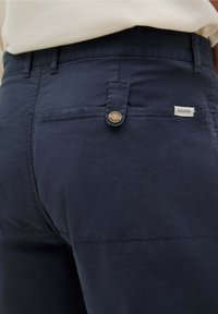 Pantalon bleu marine fabriqué dans un tissu lisse, avec une fermeture à bouton unique et une petite étiquette logo sur la poche.