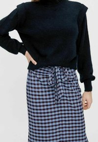 Black knit sweater with turtleneck and puffed sleeves, paired with a blue and black checkered skirt, featuring a tied waist detail.