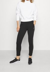 Felpa bianca con collo alto e polsini a coste, abbinata a pantaloni neri a sigaretta con strisce laterali bianche. Scarpe da ginnastica nere.
