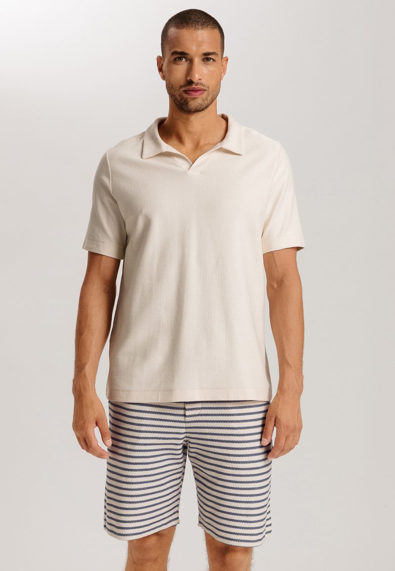 Camisa polo de manga corta beige con textura acanalada y un cuello sutil, combinada con pantalones cortos a rayas azul marino y blanca. Atuendo casual de verano.