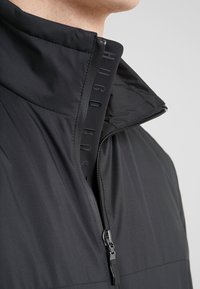 Černá zateplená bunda s vysokým límcem, předním zipem a raženým logo detailem podél zipu. Hladká textilie.