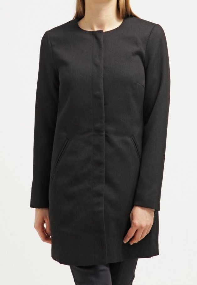 Musta, polvenpituinen takki, jossa pyöreä pääntie, pitkät hihat ja kaksi etutaskua. Kangas näyttää rakenteelliselta ja takki istuu vartalolle.