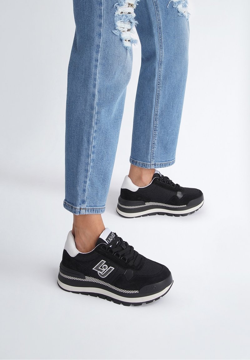 Jambes portant un jean bleu déchiré et des baskets noires avec des détails blancs, debout sur un fond uni gris clair.