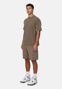 Bruine katoenen T-shirt- en shortset met witte contrasterende stiksels. Model draagt grijze en witte sneakers. Eenvoudig ontwerp, relaxte pasvorm.