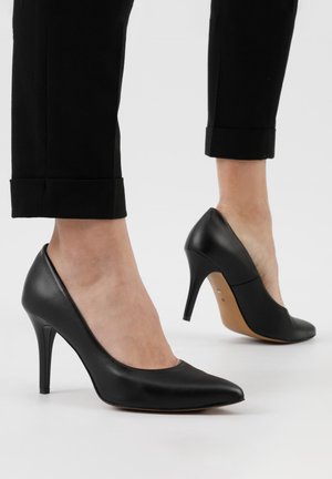 Décolleté neri in pelle con tacco alto, punta affusolata, texture liscia, finitura opaca e tacco sottile. Indossati con pantaloni neri a orlo arrotolato.