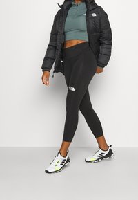 Schwarze Daunenjacke über einem türkisfarbenen Crop-Top, kombiniert mit schwarzen Leggings mit Logo und weißen Sportschuhen mit neonfarbenen Akzenten.