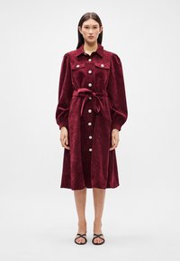 Bordó bársonyruha puffos ujjal, gombos elülső záródással és hozzá illő megkötős övvel; a gombokon virágos díszítéssel.