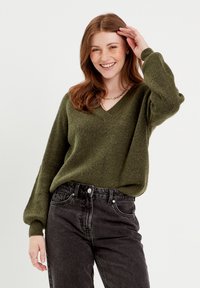 Pull en tricot vert olive avec un col en V et des manches bouffantes, associé à un jean noir taille haute avec des poches avant et une fermeture à bouton.