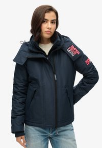 Granatowa, pikowana kurtka z kapturem i zapięciem na zamek, z naszywkami na klatce piersiowej i rękawach, bocznymi kieszeniami oraz fakturowanym wykończeniem materiału. Zawiera regulowany kaptur.