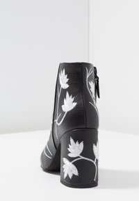 Bota de cuero negra hasta el tobillo con bordado floral blanco, cierre de cremallera, tacón apilado y textura suave. Los acentos incluyen patrones de hojas.