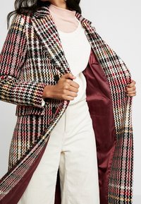 Kostkovaný kabát v červené, černé a žluté barvě, s klopovým límcem, zapínáním na knoflíky a lesklou bordó podšívkou.