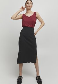 Rotes ärmelloses Oberteil mit gerafften Schultern, kombiniert mit einem knielangen schwarzen gerippten Rock mit einem seitlichen Schlitz. Schwarze Loafers mit goldenen Akzenten.