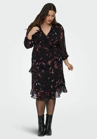 Mujer con un vestido envolventemente negro de estampado, con volantes, mangas transparentes, medias negras y botas de tobillo negras, de pie contra un fondo blanco.