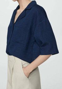 Navy blue short-sleeve shirt with a relaxed fit, lapel collar, front pocket, and textured fabric, paired with beige high-waisted trousers.