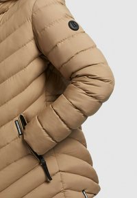 Doudoune beige avec une texture matelassée, présentant un patch de logo noir sur la manche et deux poches latérales zippées.