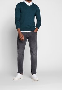 Maglione lavorato a maglia color verde acqua con scollo a V, abbinato a jeans grigi e scarpe da ginnastica bianche. Tessuto liscio e design aderente con dettagli minimi.