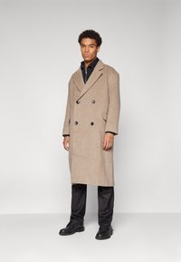 Beige doppelt gefütterter Wollmantel mit großen schwarzen Knöpfen, breiten Revers und langen Ärmeln, kombiniert mit schwarzen Hosen und Schuhen.