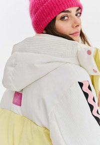Nafukovacia bunda v bielej a žltej farbe, s rebrovanou textúrou, ružovou nášivkou a čiernymi vlnitými akcentmi; dizajn kapucne s detailom gombíka.