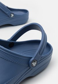 Tamancos de borracha azuis com parte superior texturizada, forma arredondada e uma alça traseira. Apresentam um botão com logotipo preto e detalhes em pontos.