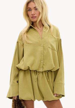 Femme blonde portant une chemise boutonnée oversize couleur olive claire et un short assorti avec cordon, tenant un accessoire en fourrure marron.