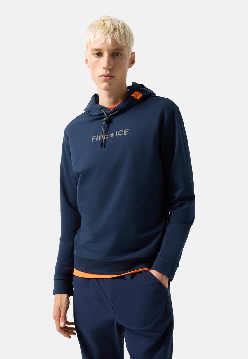 Mladý muž s krátkými blond vlasy, oblečený v námořnicky modré mikině s nápisem "FIRE + ICE" a ladících námořnicky modrých kalhotách, stojící s jednou rukou v kapse.