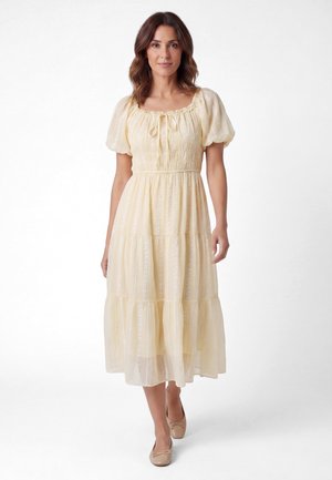 Kvinde stående iført en lysegul midi kjole med pufærmer og sløjfedetalje, kombineret med beige balletsko.