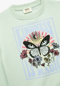 Jasnozielona bluza z okrągłym dekoltem, z grafiką motyla, kwiatowymi akcentami i napisem "ŻYCZLIWOŚĆ TO MAGIA" w wyrazistych kolorach.