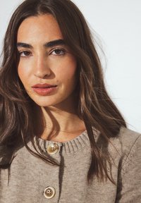 Béžs kardigāns ar ribotām apkaklītēm un lieliem, spīdīgiem pogām. Mīksta tekstūra, pieguloša forma un garas piedurknes, kas redzamas apkakles apkārtnē.