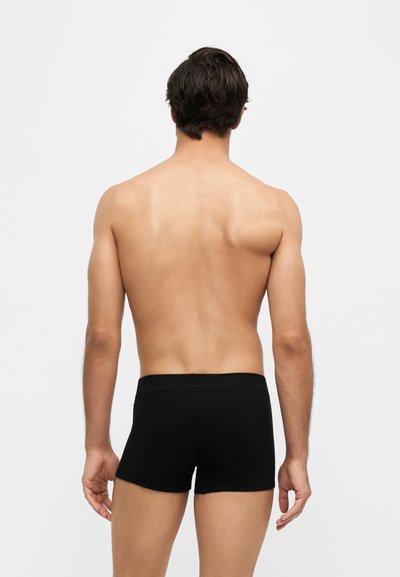 Modèle masculin debout, dos à la caméra, portant un boxer noir ajusté sur un fond clair uni.