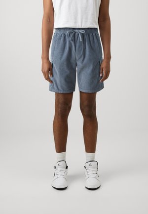 Person, der knielange blaue Cordshorts mit Kordelzug, weiße Socken, weiße High-Top-Sneaker und ein in die Hose gestecktes weißes T-Shirt trägt, steht vor einem grauen Hintergrund.