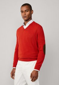 Muž ve jasně červeném svetr s V výstřihem a hnědými loketními nášivkami, pod kterým má bílou košili, stojící na pozadí v jednoduché šedé barvě.