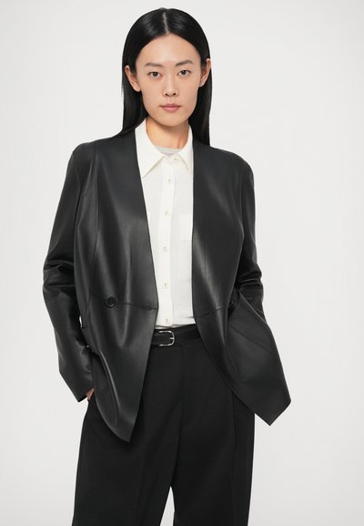 Melns ādas žakete ar strukturētu dizainu, divu pogu āķis, valkāts virs baltas krekla un melnas bikses. Gluda tekstūra un pieguļošs piegriezums.