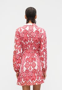 Γυναίκα με πλεξούδες, ντυμένη με κόκκινο  και λευκό φόρεμα με ζώνη και μακριά μανίκια, εμφανιζόμενη από πίσω με φόντο απλό.