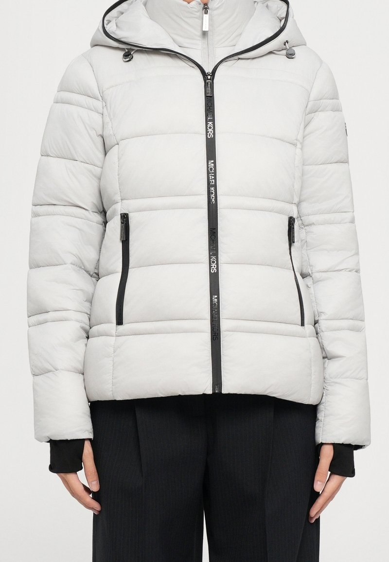 Veste matelassée gris clair avec capuche, fermeture éclair complète à l'avant portant l'inscription "Michael Kors", poches latérales zippées, portée avec un pantalon noir.