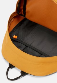 Sac à dos en tissu orange, avec une doublure intérieure noire, une ouverture zippée et une étiquette extérieure avec un logo. Texture lisse et forme arrondie.