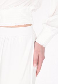 Biała, lekka bluzka z marszczonymi mankietami i pasem, w zestawie z luźnymi białymi spodniami z boczną kieszenią i miękką teksturą.