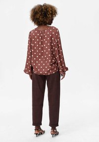 Brązowa bluzka w kropki o luźnym kroju, z długimi rękawami i plisowaniem z tyłu, w połączeniu z ciemnobrązowymi spodniami i sandałami na obcasie.