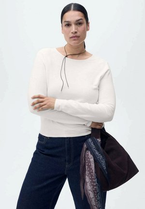 White long-sleeve top with a round neckline and black cord detail, paired with dark denim jeans and a brown bag featuring patterned scarves.