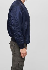 Námorně modrá bombardérka s lesklým povrchem, žebrovanými manžetami a lemem, zipovým zapínáním a boční kapsou. V kombinaci s olivově zelenými cargo kalhotami.