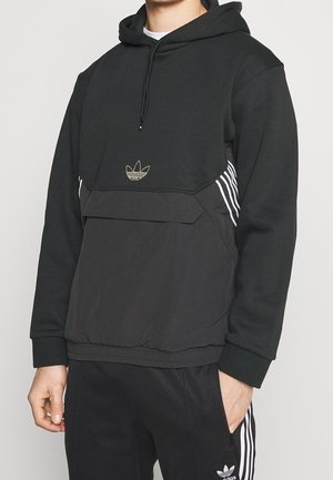 Hombre con sudadera negra Adidas con logo dorado y pantalones deportivos negros Adidas a juego con franjas blancas, con la mano en el bolsillo, sobre fondo neutro.