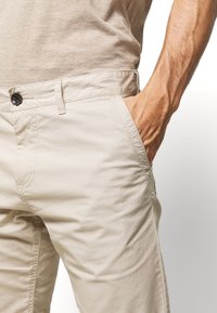 Prawa ręka spoczywa w przedniej kieszeni beżowych spodni, które noszone są z beżową, w prążki koszulą widoczną w pasie.