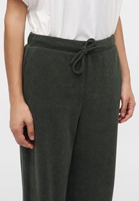 Personne portant un pantalon côtelé vert foncé avec une taille à cordon et une chemise blanche à manches courtes rentrée.