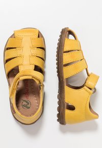 Žluté kožené sandály s otevřeným designem, které mají několik řemínků a suchý zip na kotníku. Texturovaná gumová podrážka s gripem.