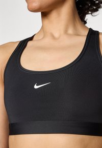 Crni sportski grudnjak od glatke tkanine, s okruglim izrezom i bijelim Nike logotipom na sredini. Neizolirana izrada, elegantan završetak.