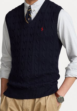 Mann trägt eine marineblaue Strickweste über einem gestreiften Hemd mit hochgekrempelten Ärmeln, eine gestreifte Krawatte mit Clip, beige Hose und eine Armbanduhr mit grünem Zifferblatt und braunem Lederarmband.