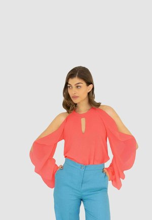Blusa in chiffon corallo con design a spalla scoperta e dettaglio a occhiello, abbinata a pantaloni azzurri. Le maniche fluide creano una silhouette morbida.
