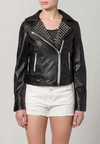 Frau in einer schwarzen Lederbikerjacke mit silbernen Nieten am Kragen und weißen Jeansshorts mit fransigem Saum.