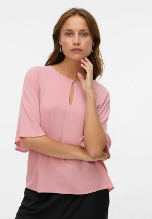 Donna con capelli castano chiaro che indossa una blusa rosa con scollatura a chiave e pantaloni neri, in posa con una mano vicino al viso.