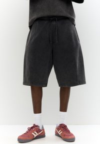 Pantaloni shorts in denim nero con una cintura elastica e cordoncino, caratterizzati da una vestibilità ampia e un design che arriva fino al ginocchio, abbinati a sneaker rosse e calze bianche.