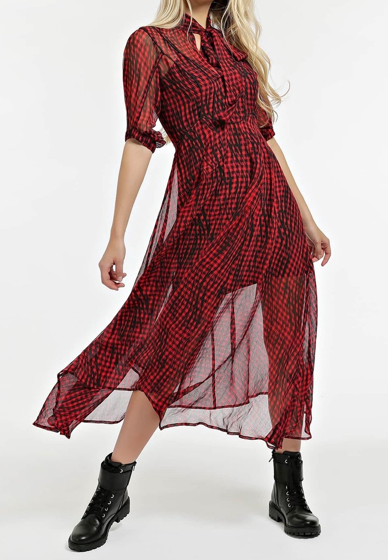 Robe maxi en tulle à carreaux rouge et noir avec des manches bouffantes et un col à nouer, présentant un design fluide et des fentes sur les côtés. Bottines noires.